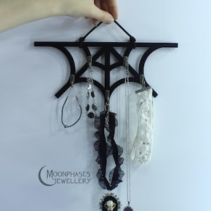May include: A black wall-mounted jewelry organizer shaped like a spider web with four metal hooks. The organizer displays various necklaces, a black ruffled choker with a skull pendant, and a white lace band. The text Moonphases Jewellery is visible.