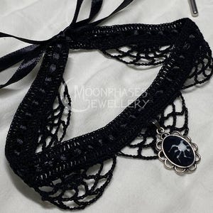 May include: Black lace choker with a silver pendant featuring a black and white image of a spider. The choker has a black satin ribbon tie.