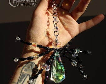 Puede incluir: Un adorno con forma de araña que presenta un gran cuerpo de cristal iridiscente en forma de lágrima, patas de cuentas facetadas negras y transparentes, y una cadena plateada. El texto Moonphases Jewellery y el número 1 son visibles.