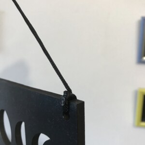 May include: A black rectangular plaque with cut-out shapes, suspended by a thin black cord threaded through a hole in the top corner. The plaque has a matte finish and is shown from a side angle, highlighting the thickness of the material.