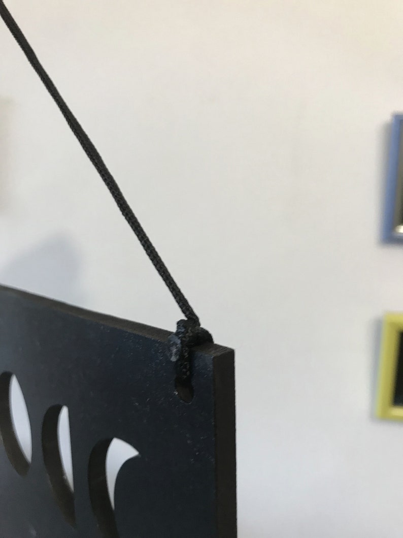 May include: A black rectangular plaque with cut-out shapes, suspended by a thin black cord threaded through a hole in the top corner. The plaque has a matte finish and is shown from a side angle, highlighting the thickness of the material.