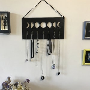 May include: A rectangular black wall-mounted jewelry organizer featuring a cutout moon phase design along the top edge. It includes seven silver-toned metal hooks for hanging necklaces, with a black cord attached to the top for wall suspension.
