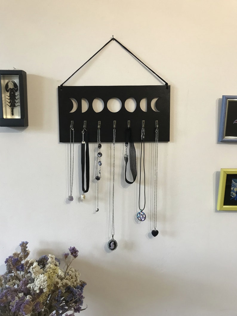 May include: A rectangular black wall-mounted jewelry organizer featuring a cutout moon phase design along the top edge. It includes seven silver-toned metal hooks for hanging necklaces, with a black cord attached to the top for wall suspension.
