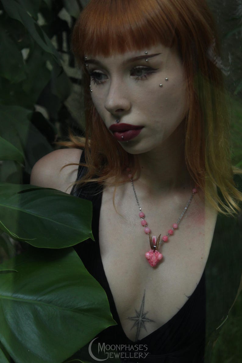 Pode incluir: Colar de corrente prateada com um pendente rosa &uacute;nico, possivelmente uma flor ou criatura. O colar tem contas rosas e &eacute; usado sobre um top preto. O fundo &eacute; folhagem verde.
