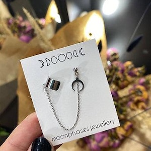 Puede incluir: Juego de pendientes y puño de oreja plateados con diseño de media luna, conectados por una delicada cadena. El conjunto se muestra en una tarjeta blanca con el texto "moonphases.jewellery" y gráficos de luna.