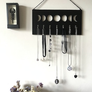 May include: A black rectangular wall-mounted jewelry organizer featuring a cutout moon phase design at the top and seven silver hooks holding various necklaces and chokers, accompanied by a small black framed display case containing a scorpion.