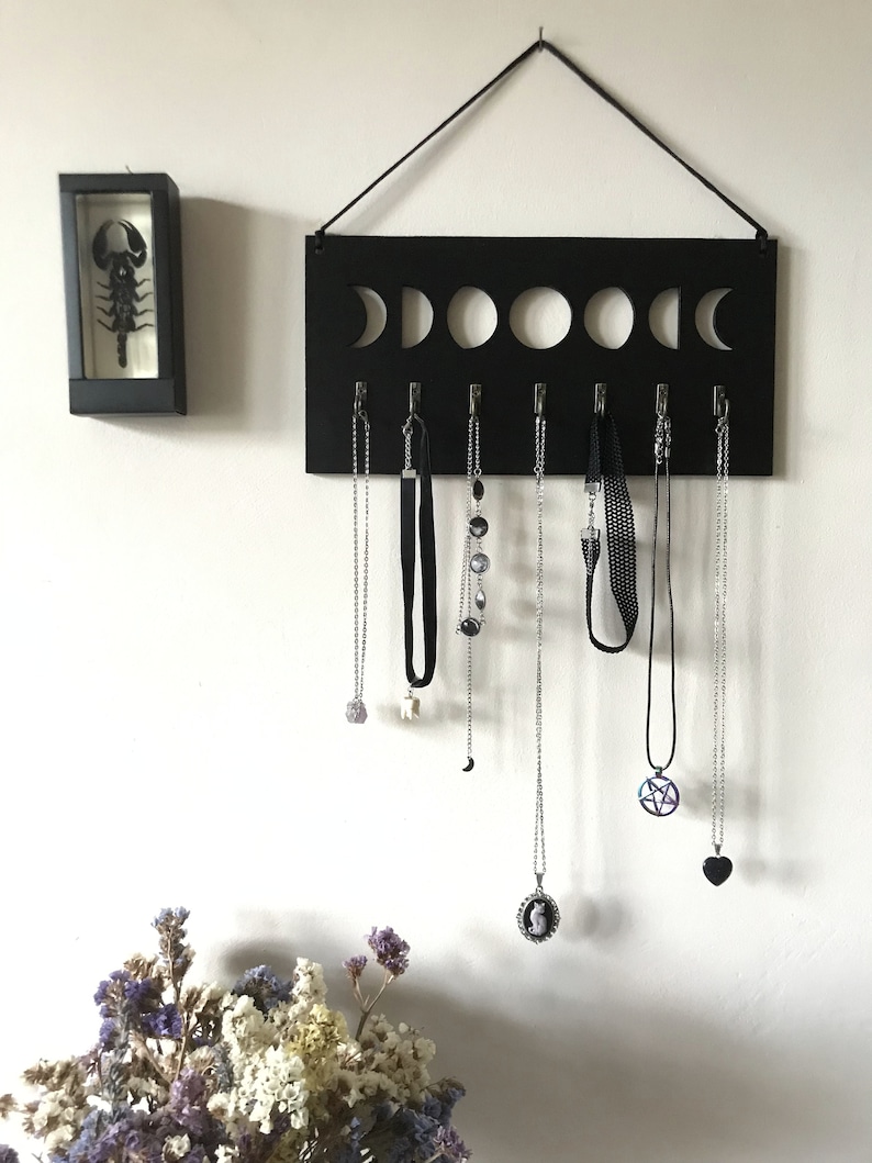 May include: A black rectangular wall-mounted jewelry organizer featuring a cutout moon phase design at the top and seven silver hooks holding various necklaces and chokers, accompanied by a small black framed display case containing a scorpion.