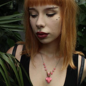 Pode incluir: Um colar rosa e prateado com um pingente de coelho. O pingente tem um design em forma de cora&ccedil;&atilde;o com um rosto de coelho. O colar &eacute; usado sobre um top preto. A imagem tamb&eacute;m mostra o texto "MOONPHASES JEWELLERY".