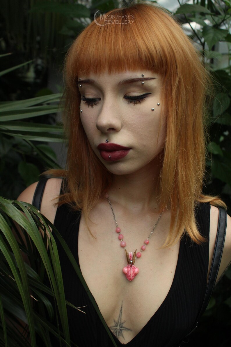 Pode incluir: Um colar rosa e prateado com um pingente de coelho. O pingente tem um design em forma de cora&ccedil;&atilde;o com um rosto de coelho. O colar &eacute; usado sobre um top preto. A imagem tamb&eacute;m mostra o texto "MOONPHASES JEWELLERY".