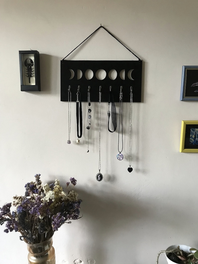 May include: A rectangular black wall-mounted jewelry organizer featuring a cutout moon phase design along the top edge and seven silver-toned metal hooks. Several necklaces with various pendants and chains are hanging from the hooks on the display.