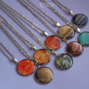 Puede incluir: Una colección de collares plateados con colgantes redondos que muestran imágenes del Sol y planetas como Mercurio, Venus, Tierra, Marte, Júpiter, Saturno, Urano y Neptuno. Cada colgante está etiquetado con su nombre en texto blanco.