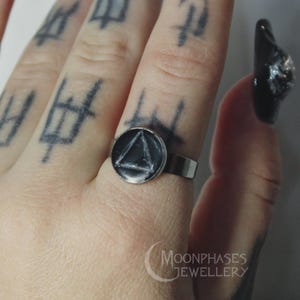 May include: A silver ring with a black circular face featuring a white triangle design. The ring is on a finger with tattoos. The Moonphases Jewellery logo is visible in the lower right corner.