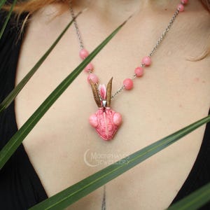 Pode incluir: Colar com pingente rosa em forma de inseto, corrente prateada e contas rosas. O pingente tem antenas marrons e um detalhe floral. O colar &eacute; exibido sobre um top preto, com folhas verdes em primeiro plano.