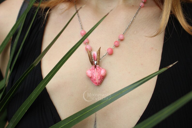 Pode incluir: Colar com pingente rosa em forma de inseto, corrente prateada e contas rosas. O pingente tem antenas marrons e um detalhe floral. O colar &eacute; exibido sobre um top preto, com folhas verdes em primeiro plano.