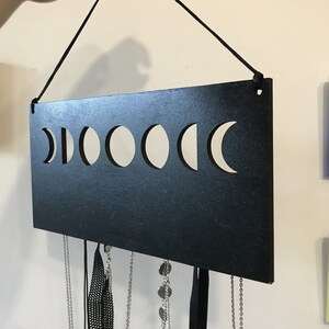 May include: A rectangular black wall-mounted jewelry organizer featuring a horizontal cutout design of the moon phases. The piece is suspended by a black cord and includes several silver-toned metal chains and black ribbons hanging from the bottom edge.