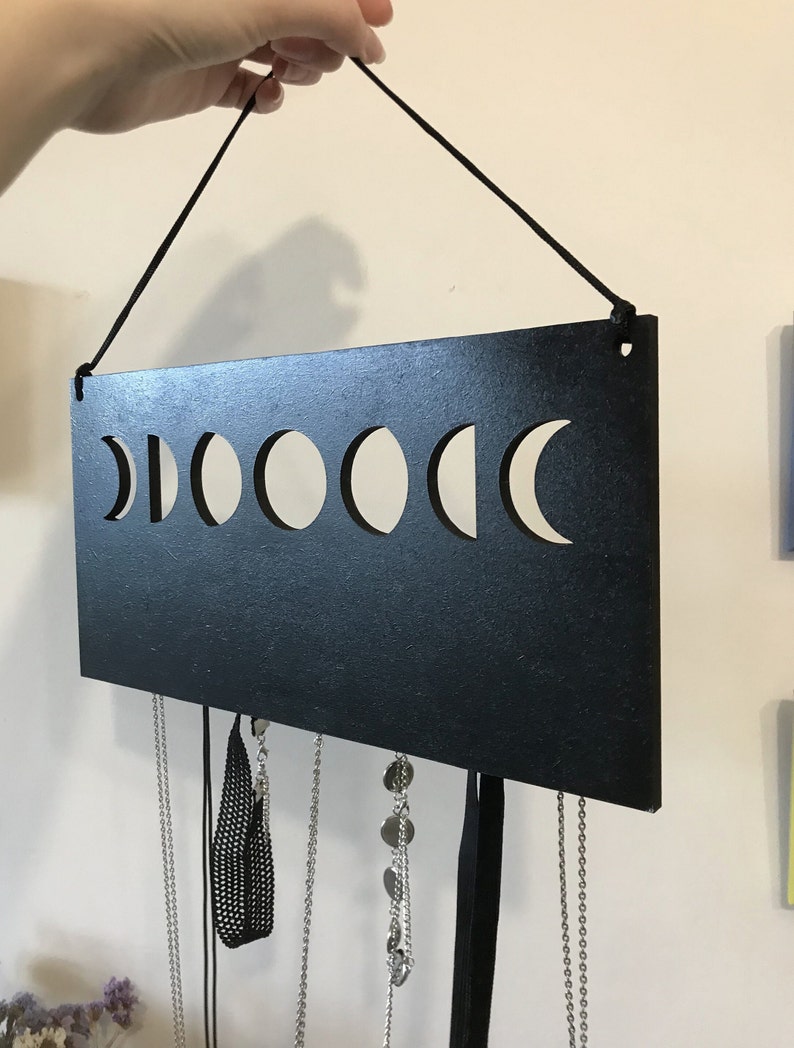 May include: A rectangular black wall-mounted jewelry organizer featuring a horizontal cutout design of the moon phases. The piece is suspended by a black cord and includes several silver-toned metal chains and black ribbons hanging from the bottom edge.