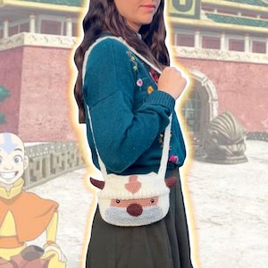 May include: A white crocheted shoulder bag shaped like Appa, the flying bison from Avatar: The Last Airbender. The bag has brown horns, a pink nose, and a gray face.