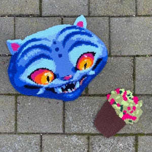 Peut inclure: Coussin en crochet en forme de chat bleu, avec des détails orange et rose. Un petit cupcake en crochet, marron, vert et rose, est également visible. Les objets sont posés sur une surface en briques.