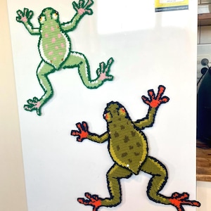 May include: Two handmade frog-shaped magnets. One is light green with pink spots and white trim, the other is olive green with orange feet and a black outline. Both are attached to a white refrigerator door.
