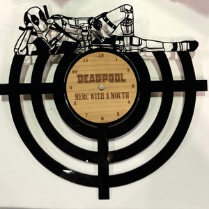 Puede incluir: Silueta en blanco y negro de Deadpool tumbado sobre un reloj de vinilo negro con una cara de madera. La esfera del reloj tiene las palabras "DEADPOOL" y "MERC WITH A MOUTH" impresas en ella.