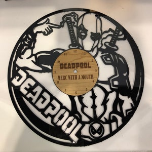 Puede incluir: Reloj de vinilo negro en forma de disco con una silueta de Deadpool con dos espadas. La esfera del reloj es un círculo de madera con las palabras "DEADPOOL MERC WITH A MOUTH" y los números del 1 al 12.