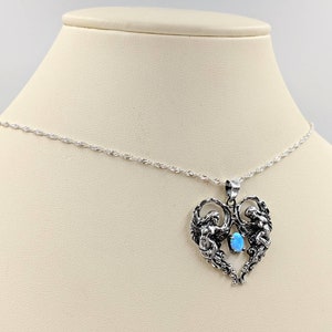 May include: A silver necklace with a heart-shaped pendant featuring two angels holding a blue opal gemstone.