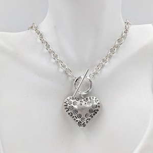 Può includere: Una collana d'argento con chiusura a scatto e un grande ciondolo a forma di cuore decorato. Il ciondolo a forma di cuore ha una superficie testurizzata con un motivo ripetitivo di piccoli cerchi.