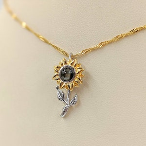 May include: A gold chain necklace with a sunflower pendant. The sunflower is made of gold and silver metal and has a dark gray gemstone in the center.