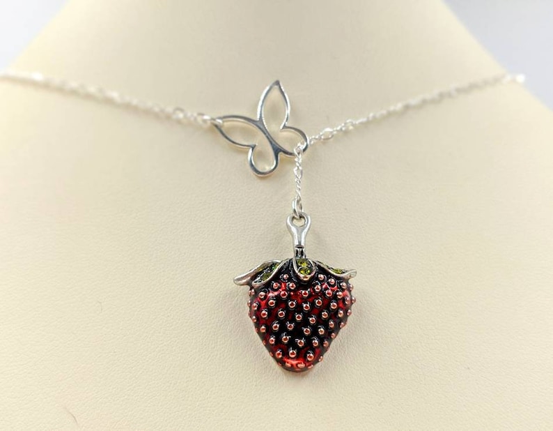 Peut inclure: Un collier en cha&icirc;ne argent avec un pendentif en forme de fraise et un pendentif en forme de papillon. La fraise est rouge avec des feuilles vertes et des graines noires. Le papillon est argent&eacute; et a les ailes d&eacute;ploy&eacute;es.