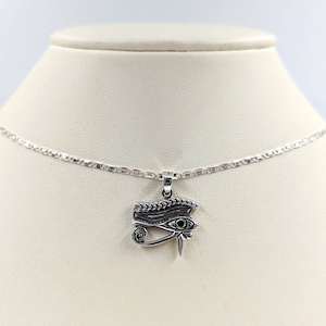 Puede incluir: Un collar de cadena de plata con un colgante de plata que presenta un diseño intrincado del Ojo de Horus, un antiguo símbolo egipcio de protección y buena salud.