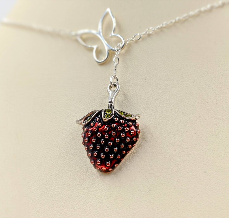 Peut inclure: Un collier en argent avec un pendentif en forme de fraise. La fraise est rouge avec des feuilles vertes et a une surface textur&eacute;e. Le collier comporte &eacute;galement un pendentif en forme de papillon argent&eacute;.