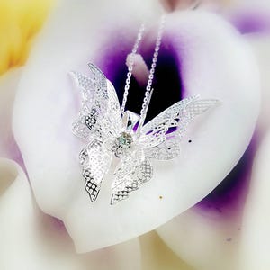 May include: A silver butterfly pendant necklace with a clear crystal in the center. The necklace is on a white and purple background.