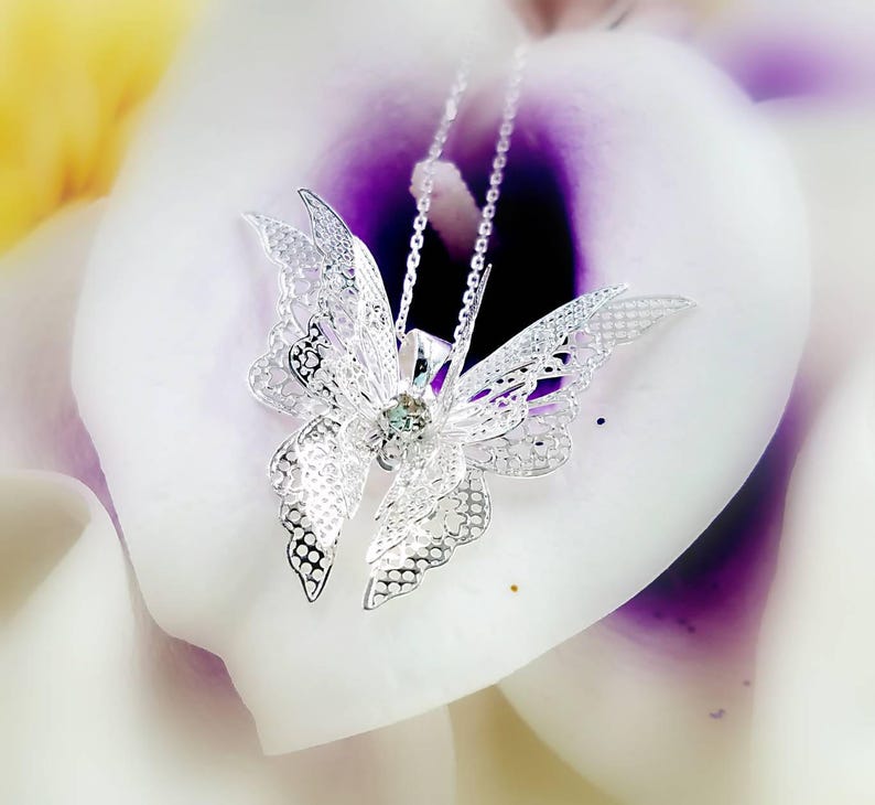 May include: A silver butterfly pendant necklace with a clear crystal in the center. The necklace is on a white and purple background.