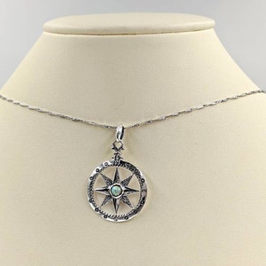 May include: A silver necklace with a compass pendant featuring a blue opal in the center. The compass has a silver star with eight points and a circle of numbers around the edge.