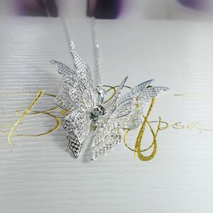 May include: Silver butterfly pendant necklace with a clear crystal in the center. The necklace is on a white background with gold lettering that says "Blossom".