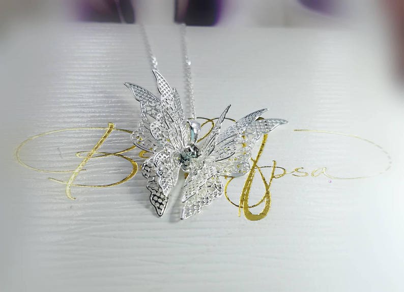 May include: Silver butterfly pendant necklace with a clear crystal in the center. The necklace is on a white background with gold lettering that says "Blossom".