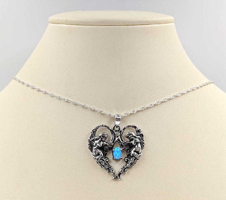 May include: A silver necklace with a heart-shaped pendant featuring two angels with wings and a blue opal gemstone in the center.