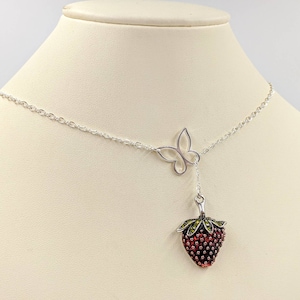 Peut inclure: Un collier en argent avec un pendentif en forme de fraise et un pendentif en forme de papillon. La fraise est rouge avec des feuilles vertes et a une finition argent&eacute;e.