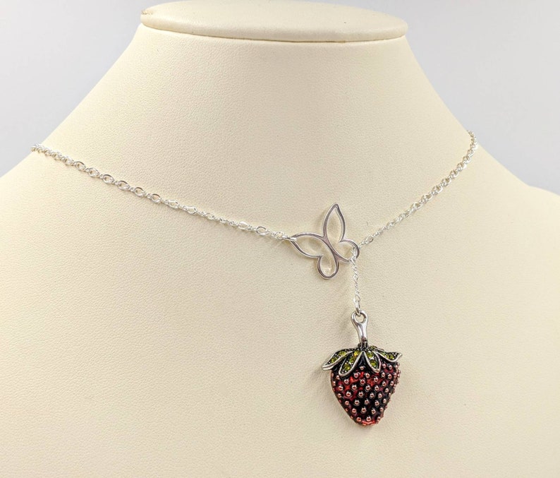 Peut inclure: Un collier en argent avec un pendentif en forme de fraise et un pendentif en forme de papillon. La fraise est rouge avec des feuilles vertes et a une finition argent&eacute;e.