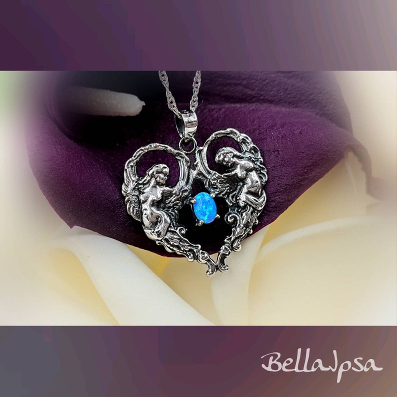 May include: A silver heart-shaped pendant with two angels holding a blue opal gemstone. The pendant is on a silver chain and is resting on a white and purple flower.