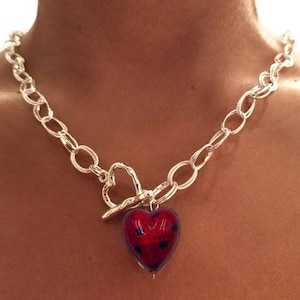 Op de afbeelding: Een zilveren ketting met een hartvormige hanger. De hanger is gemaakt van rood glas met zwarte stippen.