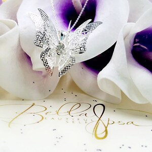 May include: A silver filigree butterfly pendant necklace with a clear crystal in the center. The necklace is displayed on a white and purple flower background.