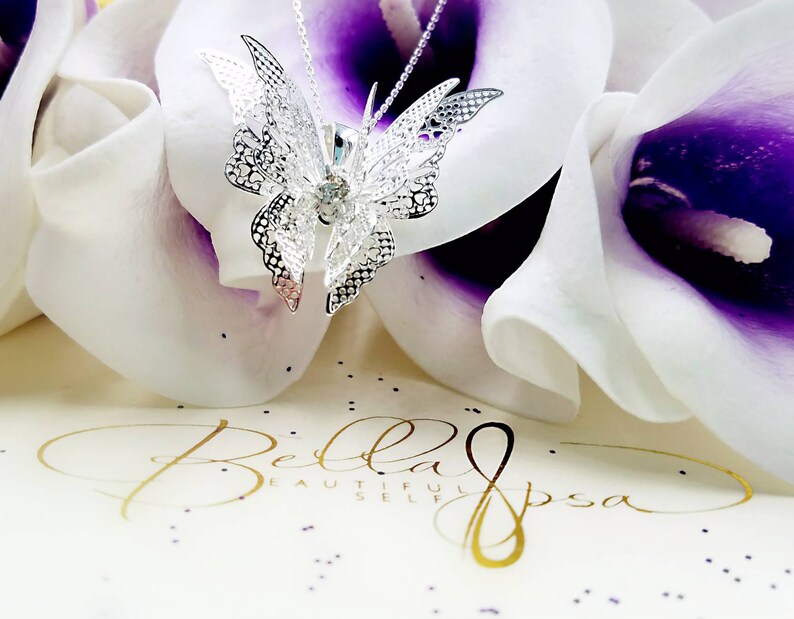 May include: A silver filigree butterfly pendant necklace with a clear crystal in the center. The necklace is displayed on a white and purple flower background.