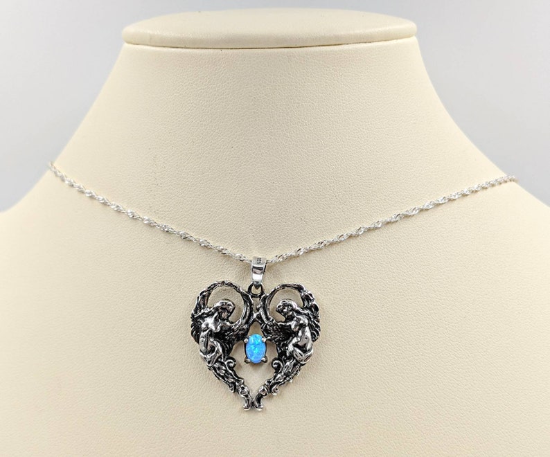 May include: A silver necklace with a heart-shaped pendant featuring two angels holding a blue opal gemstone.