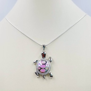 Puede incluir: Un collar de cadena de plata con un colgante de tortuga de ópalo rosa. La tortuga está detallada con acentos plateados y un acabado brillante.