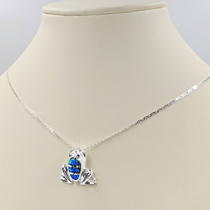 Puede incluir: Un collar de cadena de plata con un colgante de rana de ópalo azul. La rana tiene pequeños detalles rosas para los ojos.