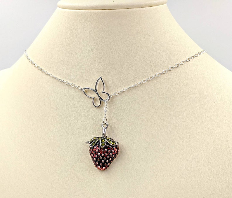 Peut inclure: Un collier en cha&icirc;ne argent avec un pendentif en forme de fraise et un pendentif en forme de papillon. La fraise est rouge avec des feuilles vertes et le papillon est argent&eacute;.