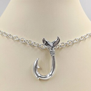 May include: A silver necklace with a fish hook pendant. The hook is detailed with a fish tail at the top.