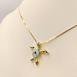 Op de afbeelding: Een goudkleurige ketting met een schildpadhanger. De hanger heeft een lichtblauw en wit opaalachtig midden. De ketting wordt gepresenteerd op een crèmekleurig oppervlak.