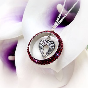 May include: A silver necklace with a pendant featuring a heart with angel wings inside a circle of red crystals.
